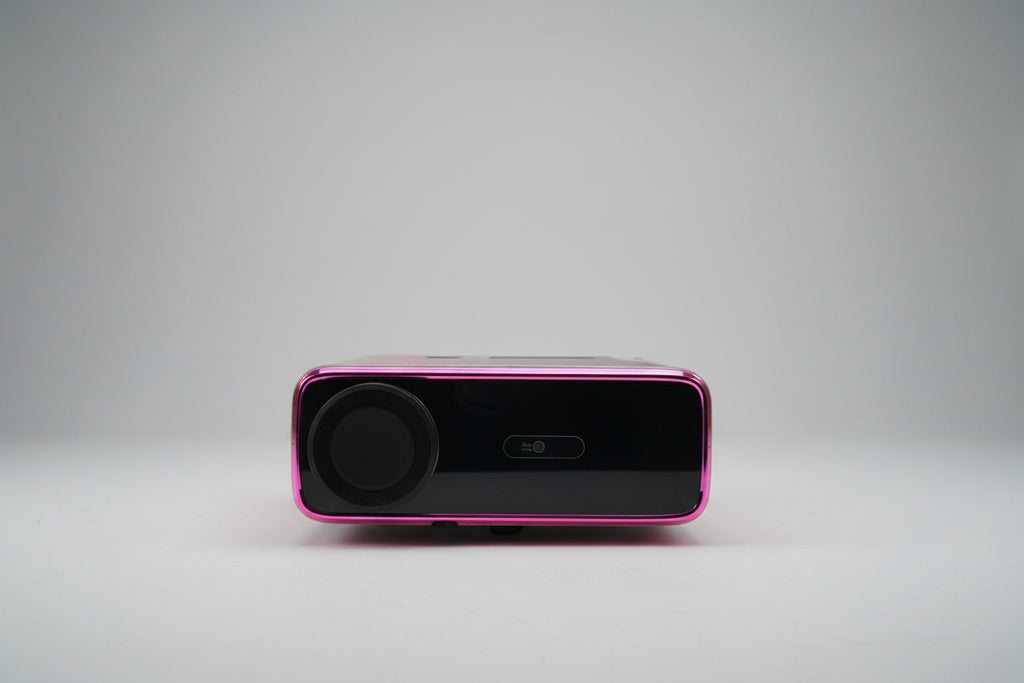 A projector with a black and pink color scheme, displaying a bright light, placed on a white stand in a room with a light source to its left.
