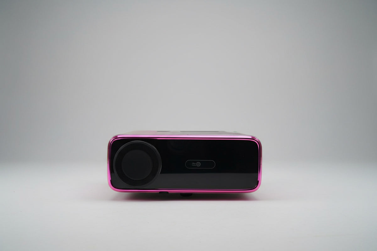 A projector with a black and pink color scheme, displaying a bright light, placed on a white stand in a room with a light source to its left.