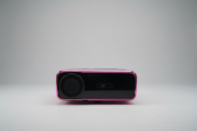 A projector with a black and pink color scheme, displaying a bright light, placed on a white stand in a room with a light source to its left.